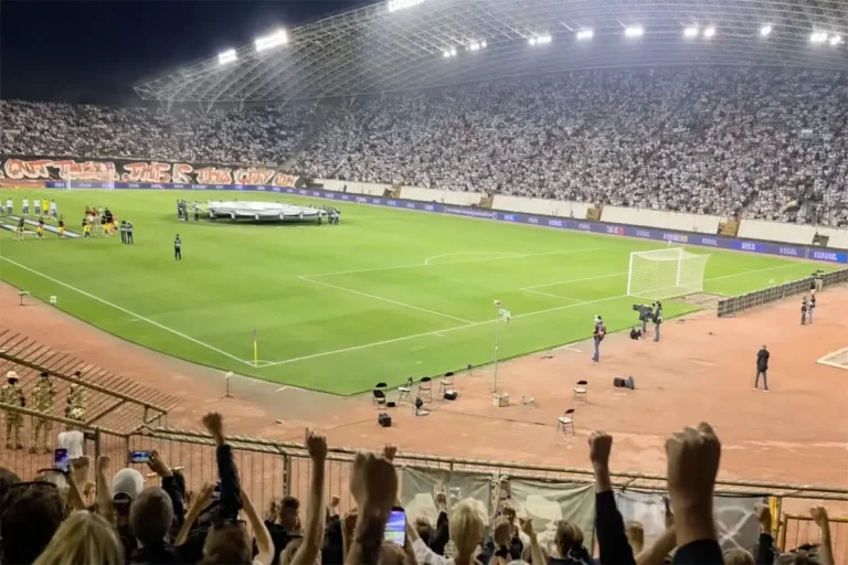 Pełny stadion w nocy podczas meczu Hajduk Split – Ajax, kibice z uniesionymi rękami na pierwszym planie, boisko i trybuny.